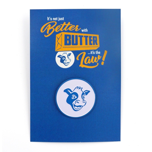 The blue and white cow head pin is situated on a blue placard that says in yellow and white  text "It's not just better with butter...it's the law."