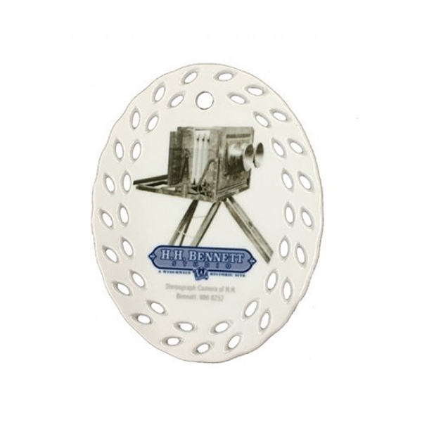 White, oval, porcelain ornament with illustration of bellows camera on a tripod