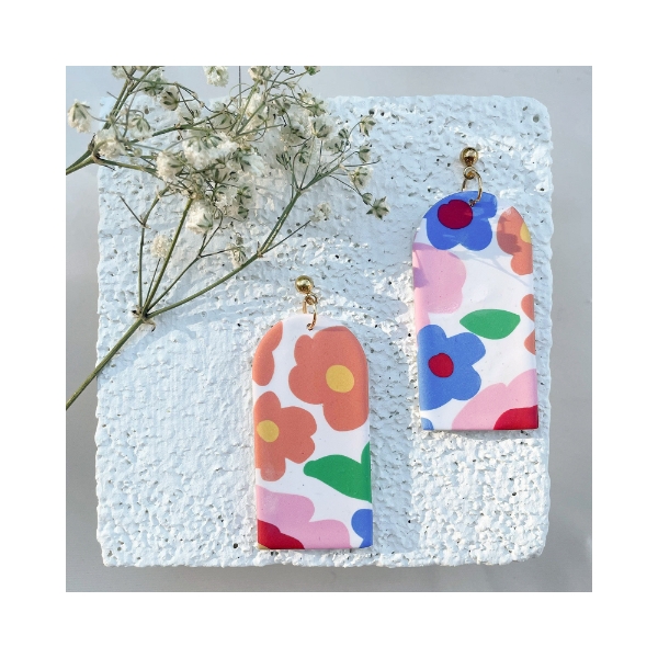Two flat floral design earrings, handmade from polymer clay. The earrings rest on a square white tile with a sprig of small wildflowers.  Colorful flower designs on a white background.