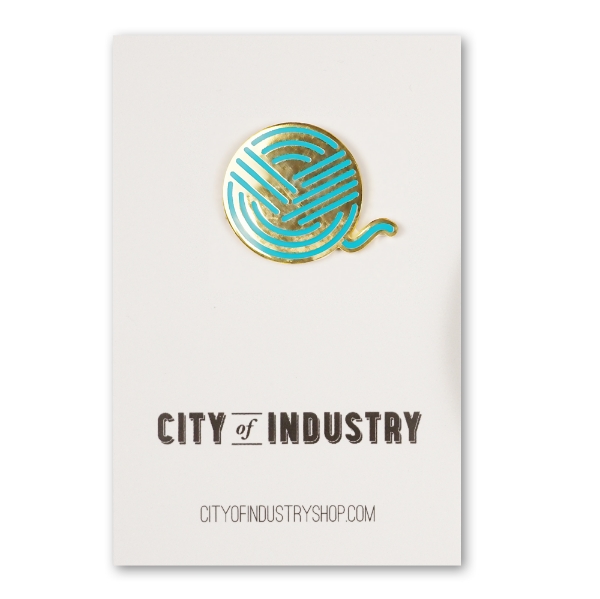 Round metal lapel pin mounted on a white card. It represents, in a stylized way, a ball of yarn. Gold chrome and blue-green enamel.