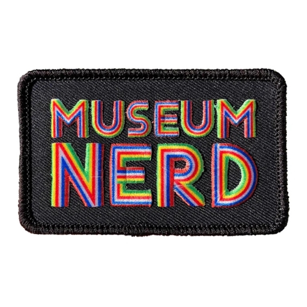 Rectangular cloth patch with rounded corners. Black background with large, striped, rainbow font that reads "Museum Nerd". 