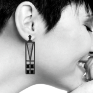 Rectangular dangle earring hanging from woman's right ear. The earring has a rectangular Frank Lloyd Wright stained glass window design with gray and black design, and it is hanging vertically. 
