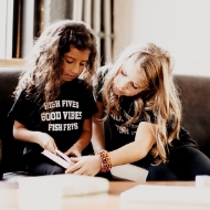 Two toddlers wearing black toddler size T-shirts that say "High Fives. Good Vibes. Fish Frys." The lettering is bold white. The two girls are sitting on a couch in a room looking at a book together.
