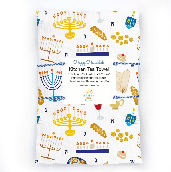 White tea towel, folded into a rectangle and bound with string, with color print design featuring renderings of dreidels, menoriahs, challah bread, and more.  
