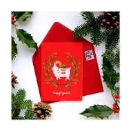 Red Yuletide card with red envelope. The red card has an illustration of a white goat encircled by a twig with green leaves. The card and evelope are surrounded by some green holly leaves and spruce twigs. 