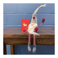 Tibbles Dangling Legs Christmas Gnome sitting on edge of a table with legs dangling. Gnome is sitting in front of a red Christmas card. 