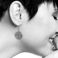 Polished metal dangle earring with perforated round shape and blue glass bead in center. Shown on woman's ear.