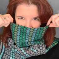 A female model wears the green, blue, and gray "Winter's Ball" scarf and pulls it up to display the honeycomb pattern. 