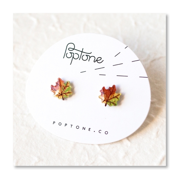 Two stud earrings in the shape of maple leaves, red and green tones. Mounted on white display card