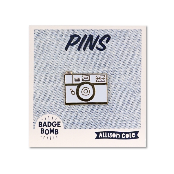 An enamel lapel pin of a white retro camera with gold outlines 